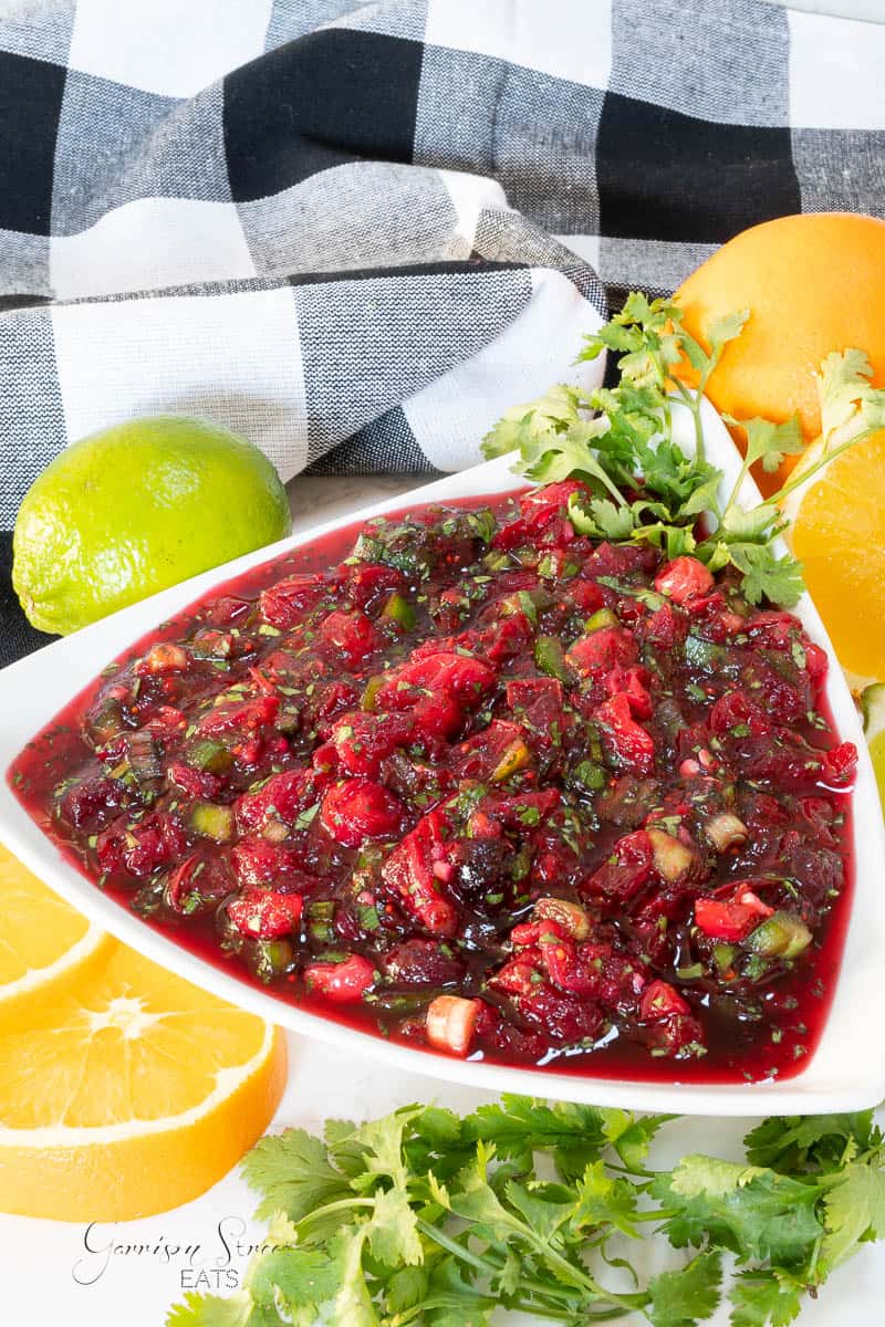 A dish of fresh cranberry salsa, is displayed on a white triangular plate. The relish includes cranberries, herbs, and diced vegetables. Surrounding the plate are vibrant lime, orange slices, cilantro, and a black-and-white checkered cloth in the background.