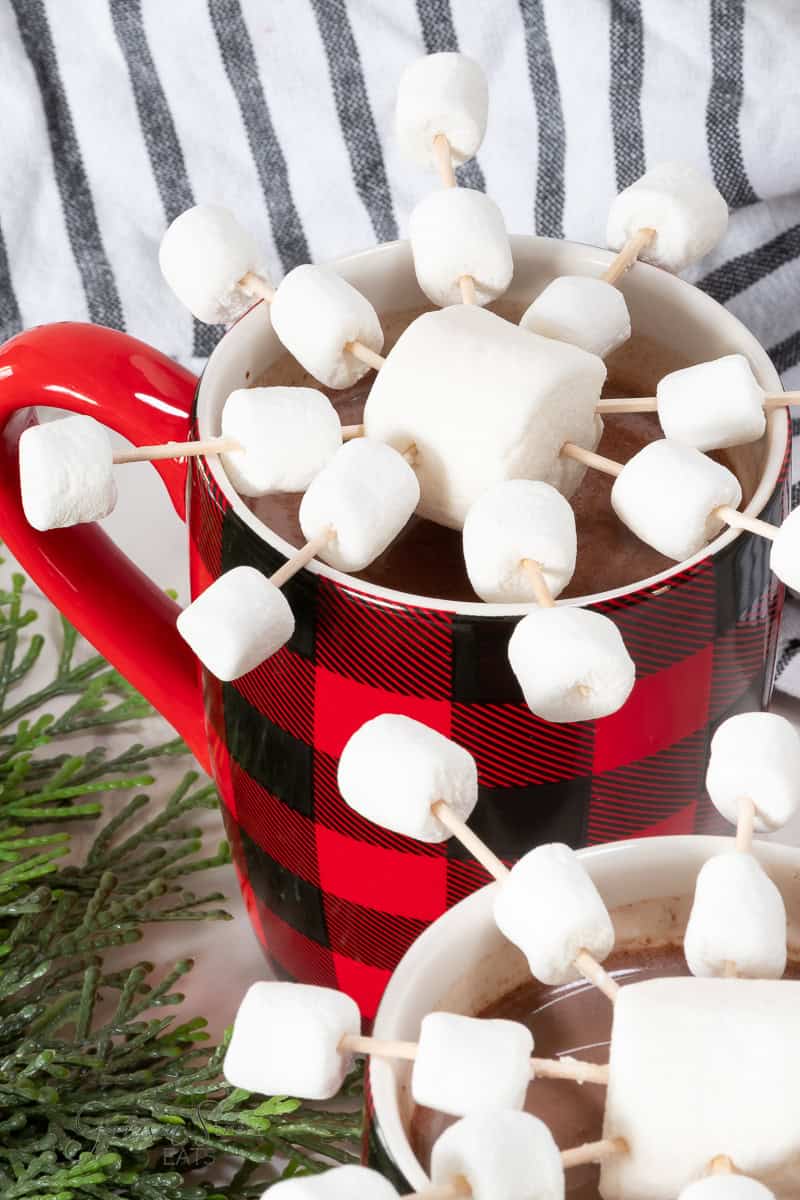 A red and black checkered mug filled with a 3 ingredient hot cocoa mix is topped with marshmallows on sticks, creating a playful starburst pattern. A smaller mug with a similar arrangement is in the foreground. A striped cloth and evergreen branches are visible in the background.