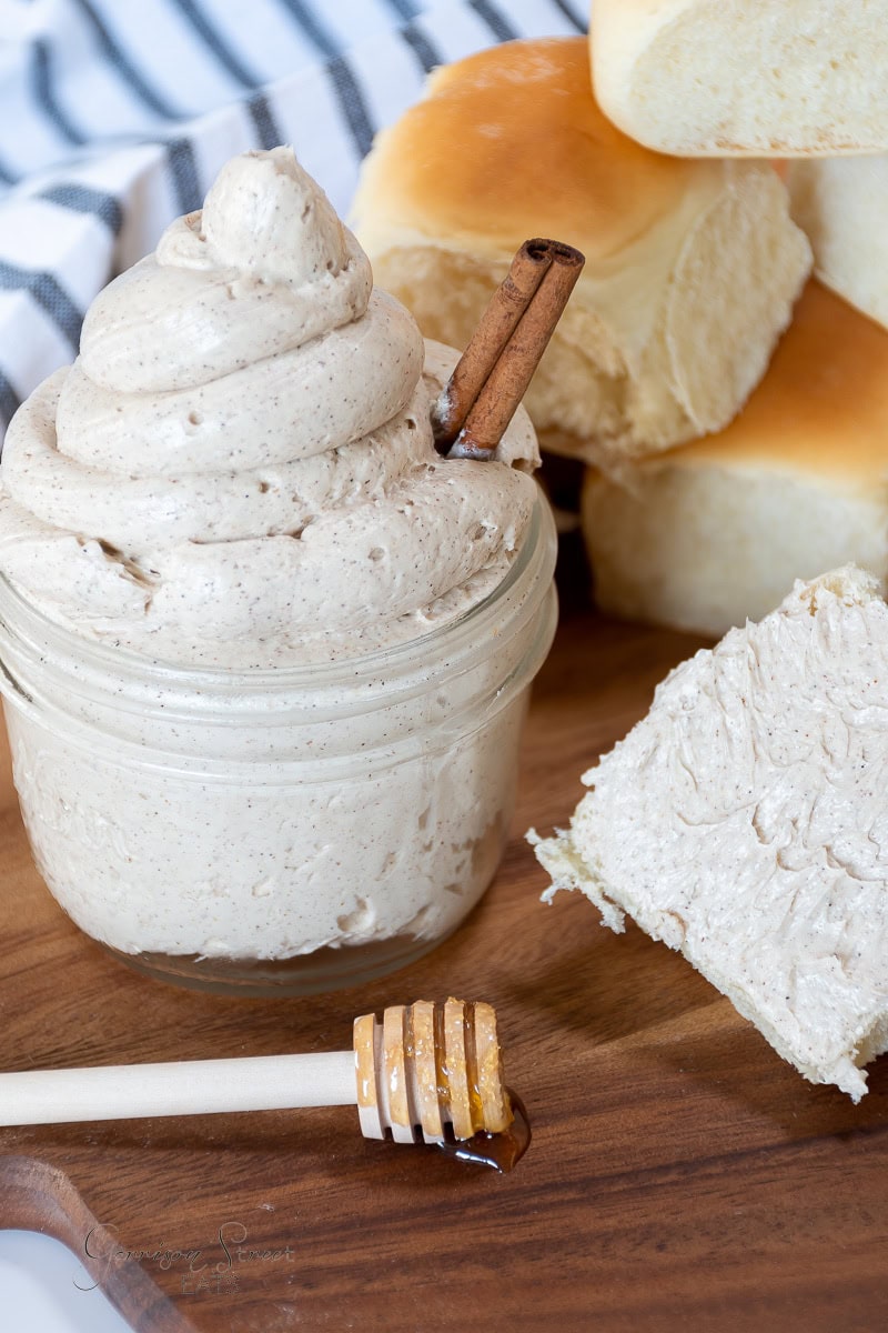 A jar of whipped cinnamon butter, is topped with a cinnamon stick and sits on a wooden board. Nearby, a bread roll slice with spread and fluffy rolls are visible. A honey dipper with honey rests in the foreground, while a striped cloth adorns the background.