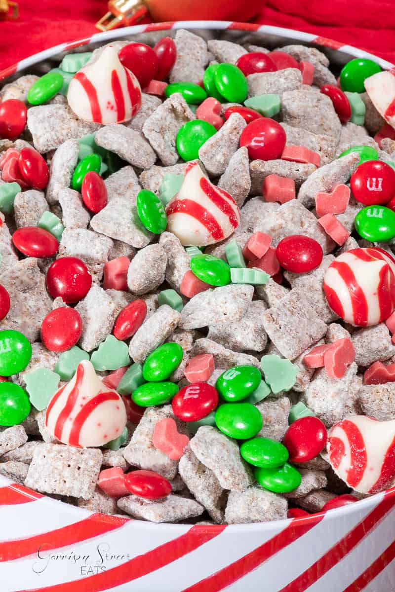 A festive mix of Christmas-themed snacks includes red and green M&M's, powdered sugar-coated cereal from a classic Christmas Muddy Buddy recipe, peppermint swirl chocolates, and red and green gummy candies in the shape of leaves and hearts, all arranged in a bowl lined with red and white striped paper.