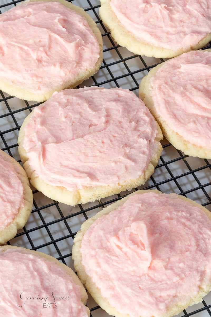 A close-up of several frosted sugar cookies sitting on a wire cooling rack. Each cookie is topped with a generous amount of pink frosting. The frosted cookies appear soft and the frosting is smoothly spread, creating a tempting display.