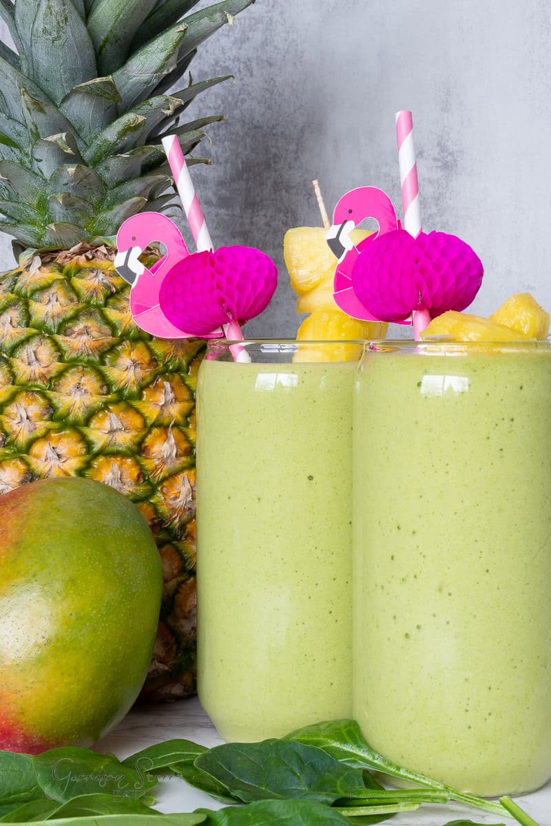 Two tall glasses of green smoothie garnished with pink flamingo straws and pineapple chunks, perfect for learning how to make a mango pineapple smoothie. A whole pineapple and a mango are in the background, with fresh spinach leaves beside the glasses. The scene has a fresh, tropical vibe.