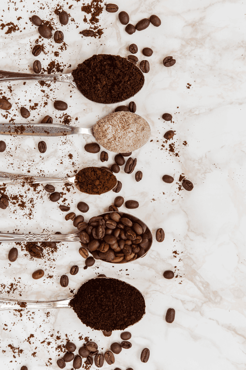 Top view of five spoons on a marble surface, each filled with different forms of coffee: whole beans, ground coffee, and powdered coffee. Coffee beans and grounds are scattered around the spoons, creating a rustic and inviting scene.