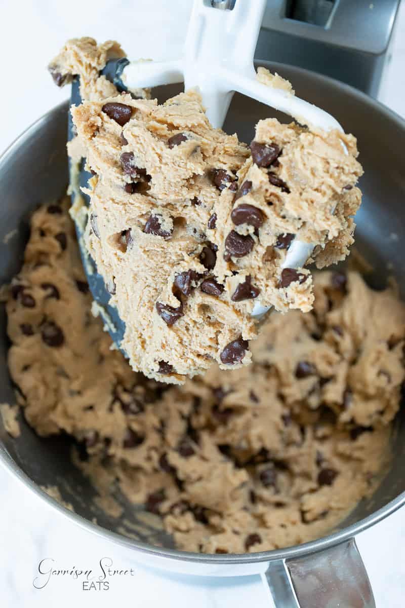 A mixing bowl filled with chocolate chip cookie dough is attached to a stand mixer. The dough, packed with chocolate chips, clings to the paddle attachment, with some dough remaining in the bowl below.