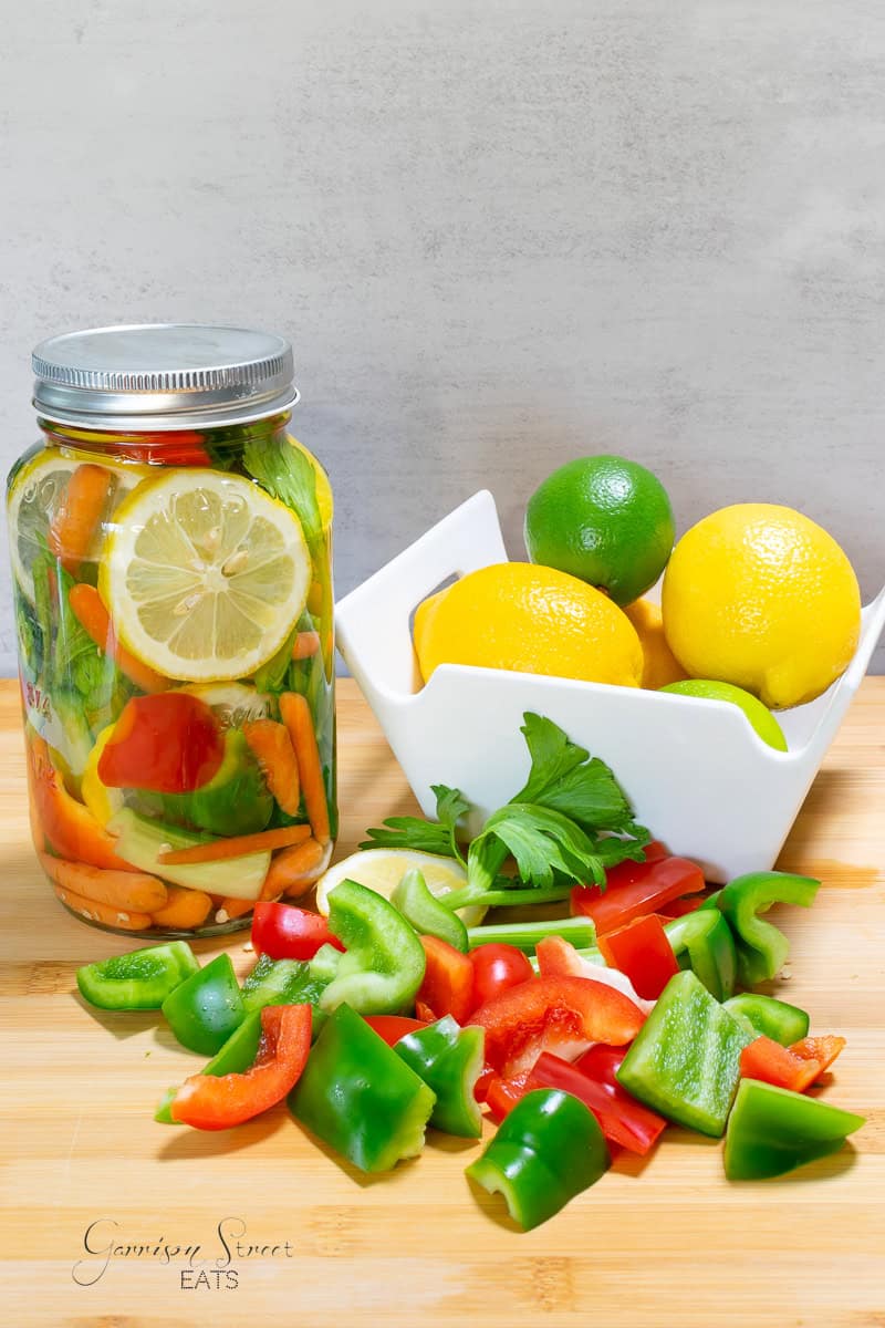 A countertop display features a jar of colorful pickled vegetables, including carrots, bell peppers, and a lemon slice. Beside it is a white bowl containing whole lemons and limes, with chopped green and red bell peppers scattered on a wooden cutting board.