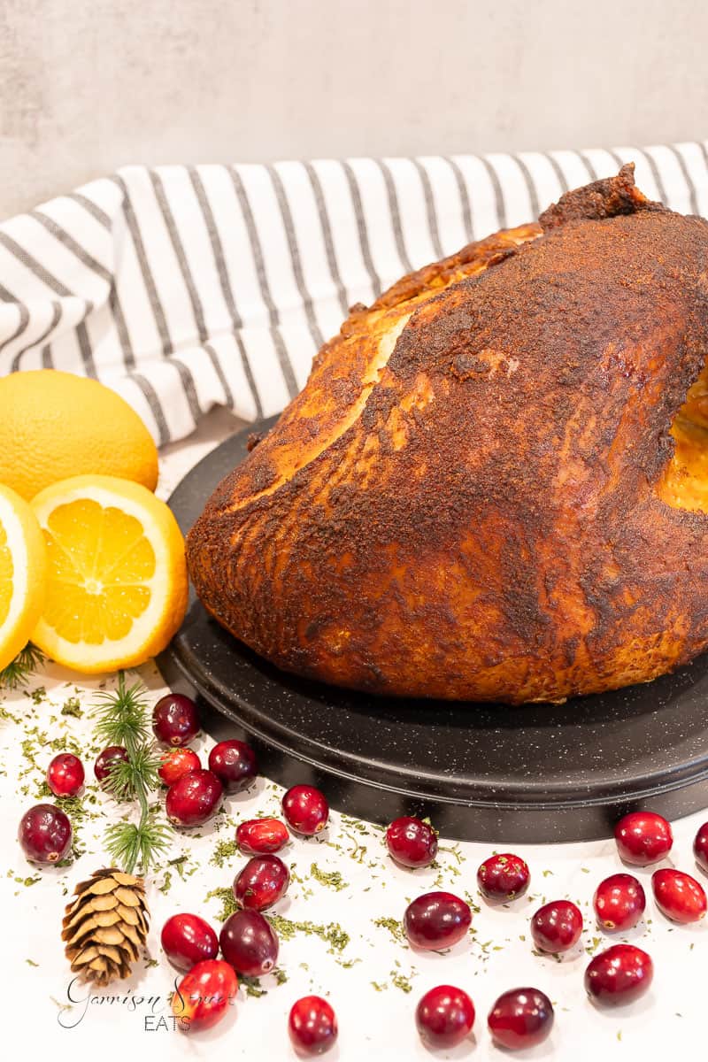 A roasted turkey breast with a crispy, seasoned skin is presented on a black platter. Sliced oranges, fresh cranberries, a small pine cone, and green herbs adorn the scene with a striped cloth in the background, adding a festive touch.