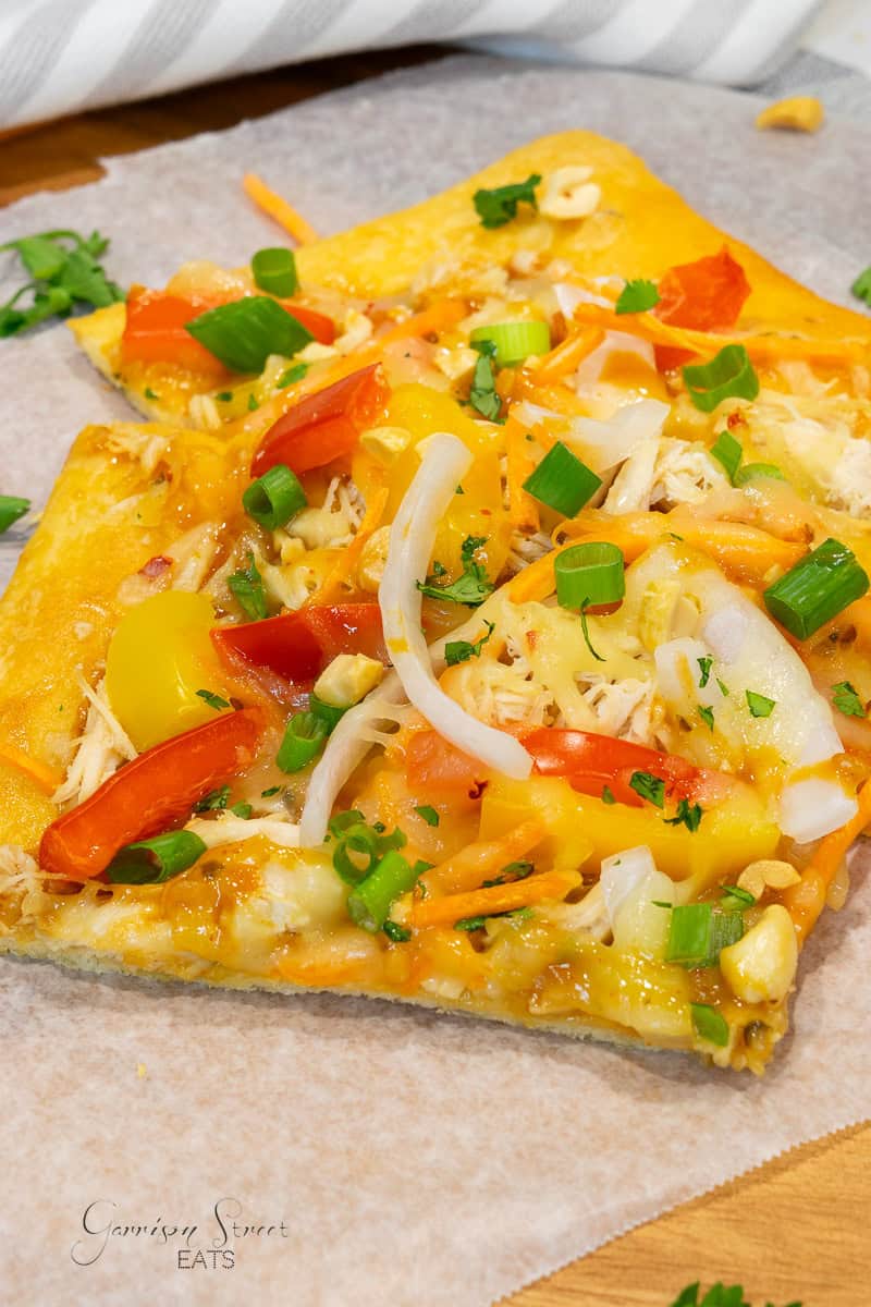 A close-up of a square slice of pizza on parchment paper, topped with shredded chicken, bell peppers, onions, green onions, and melted cheese. The crust is thin and golden brown, and there are some scattered herbs for garnish.