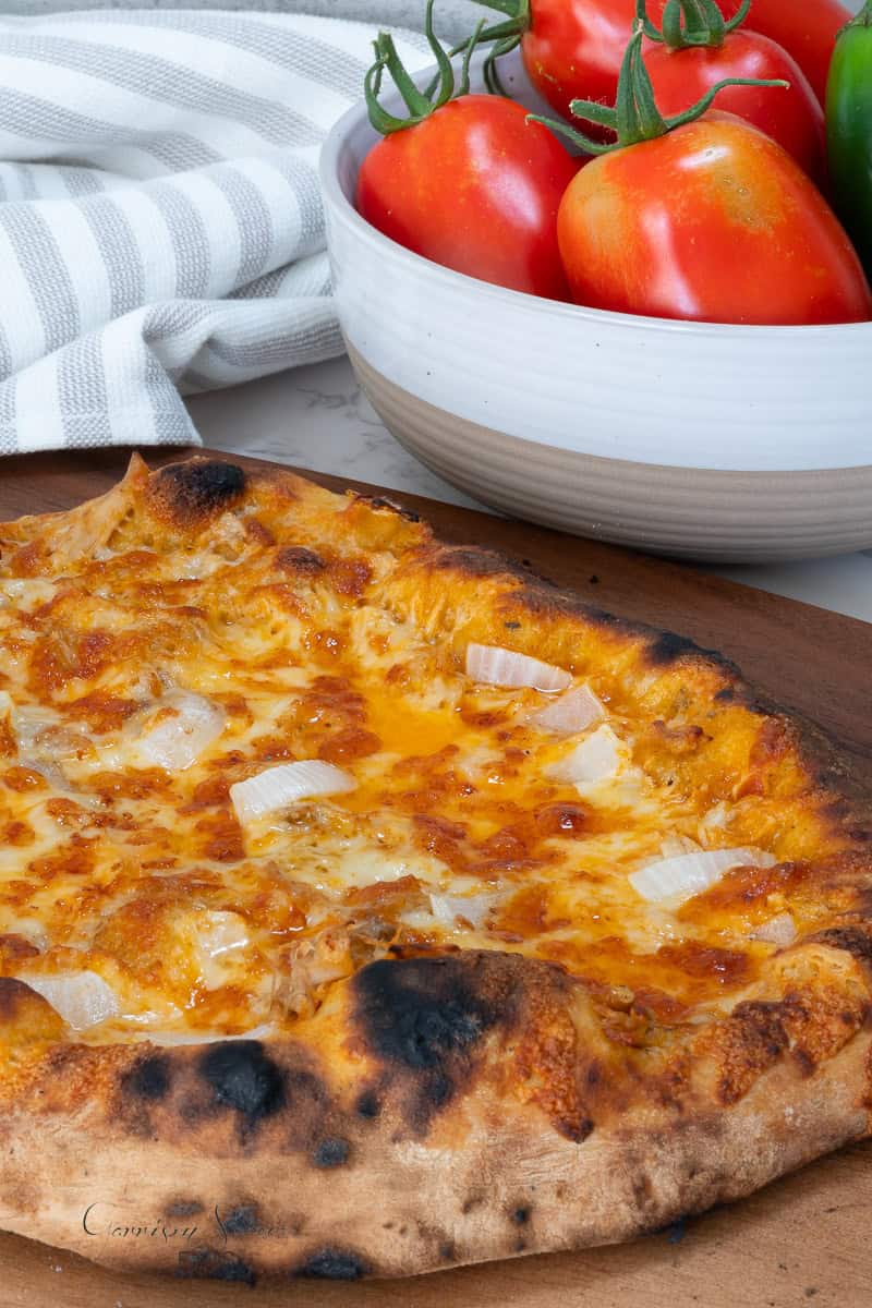 A homemade buffalo chicken pizza with a crispy, slightly charred crust, topped with melted cheese and diced onions. In the background, there is a white bowl containing fresh tomatoes and a green jalapeno. A striped dish towel is placed beside the bowl.