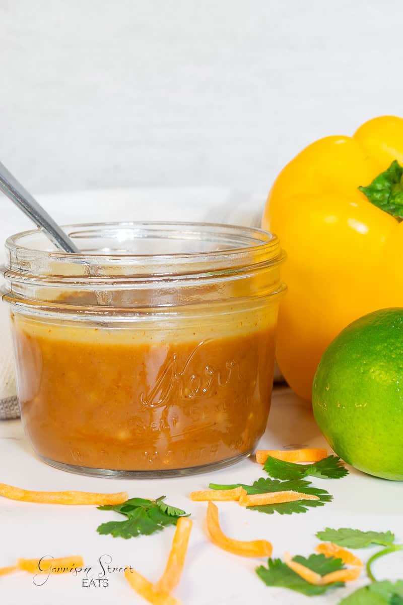 A glass jar filled with a light brown pad thai sauce sits on a white surface. A spoon is in the jar, surrounded by a yellow bell pepper, a lime, cilantro leaves, and thin orange vegetable strips. A white wall serves as the background.