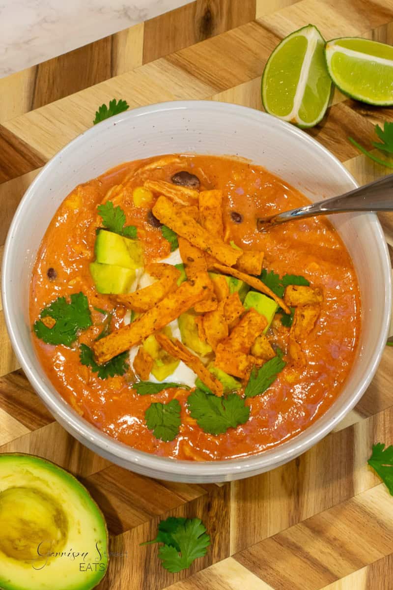 A bowl of creamy taco soup recipe with Rotel, topped with avocado chunks, tortilla strips, and fresh cilantro. Beside the bowl are lime wedges and an avocado half on a wooden cutting board. A spoon is placed in the bowl, ready for serving.