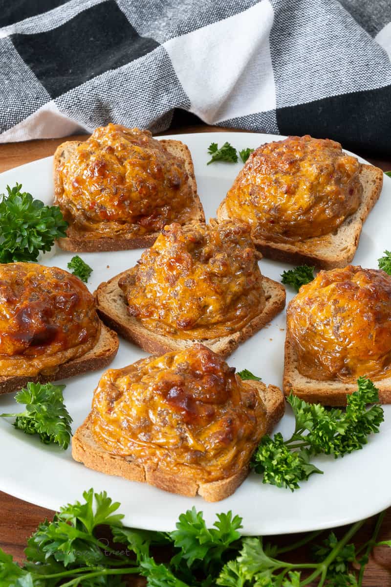 A plate of cheesy beef toast topped with a golden-brown, baked, cheesy beef mixture is garnished with fresh parsley. This delicious dish, is served on square slices of toasted bread. In the background, a black and white checkered cloth adds a rustic touch.