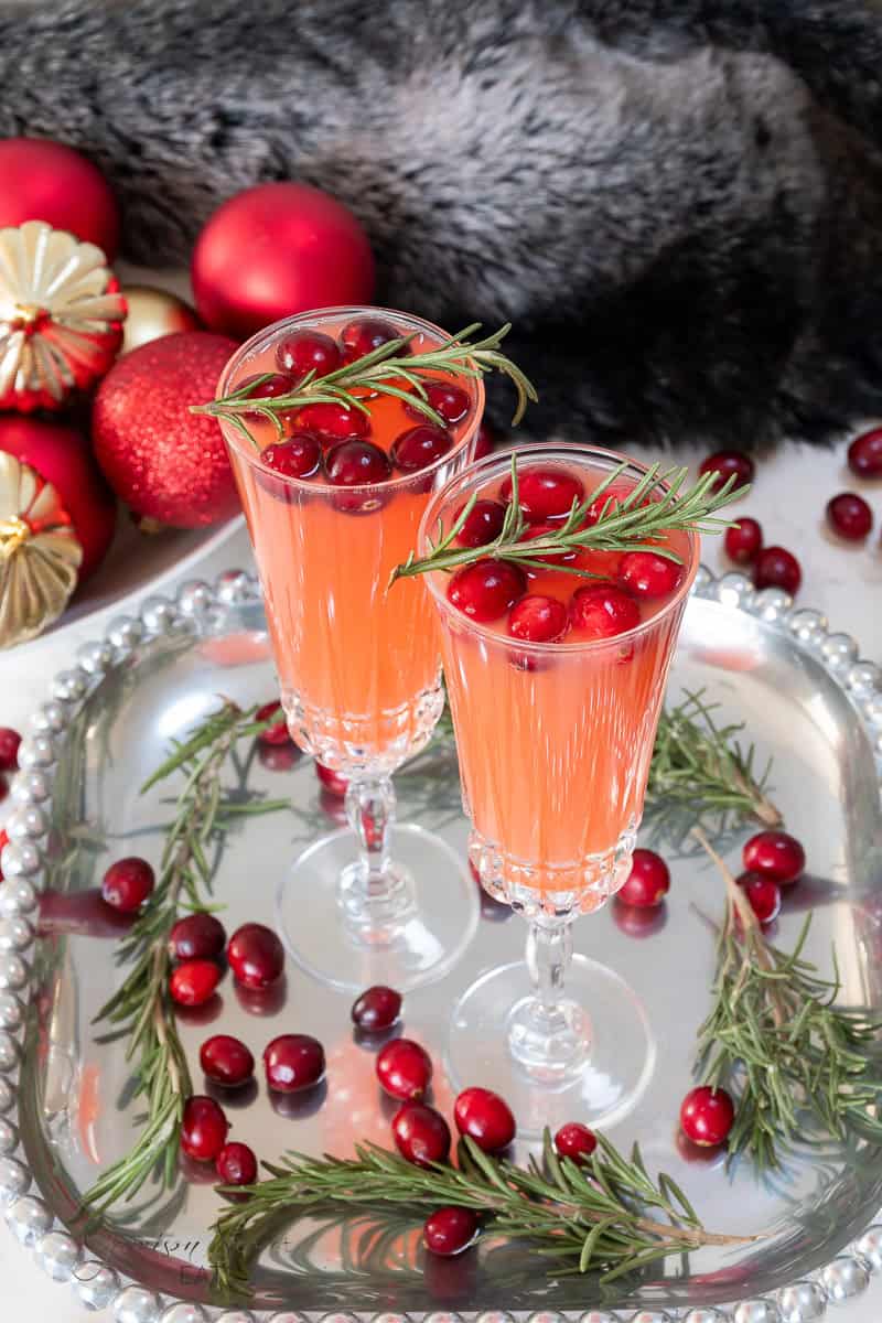 Two elegant champagne flutes filled with a festive cranberry Christmas cocktail are garnished with fresh cranberries and sprigs of rosemary. The drinks are placed on a round silver tray, which is also adorned with cranberries and rosemary. Red ornaments and a fur fabric are in the background.