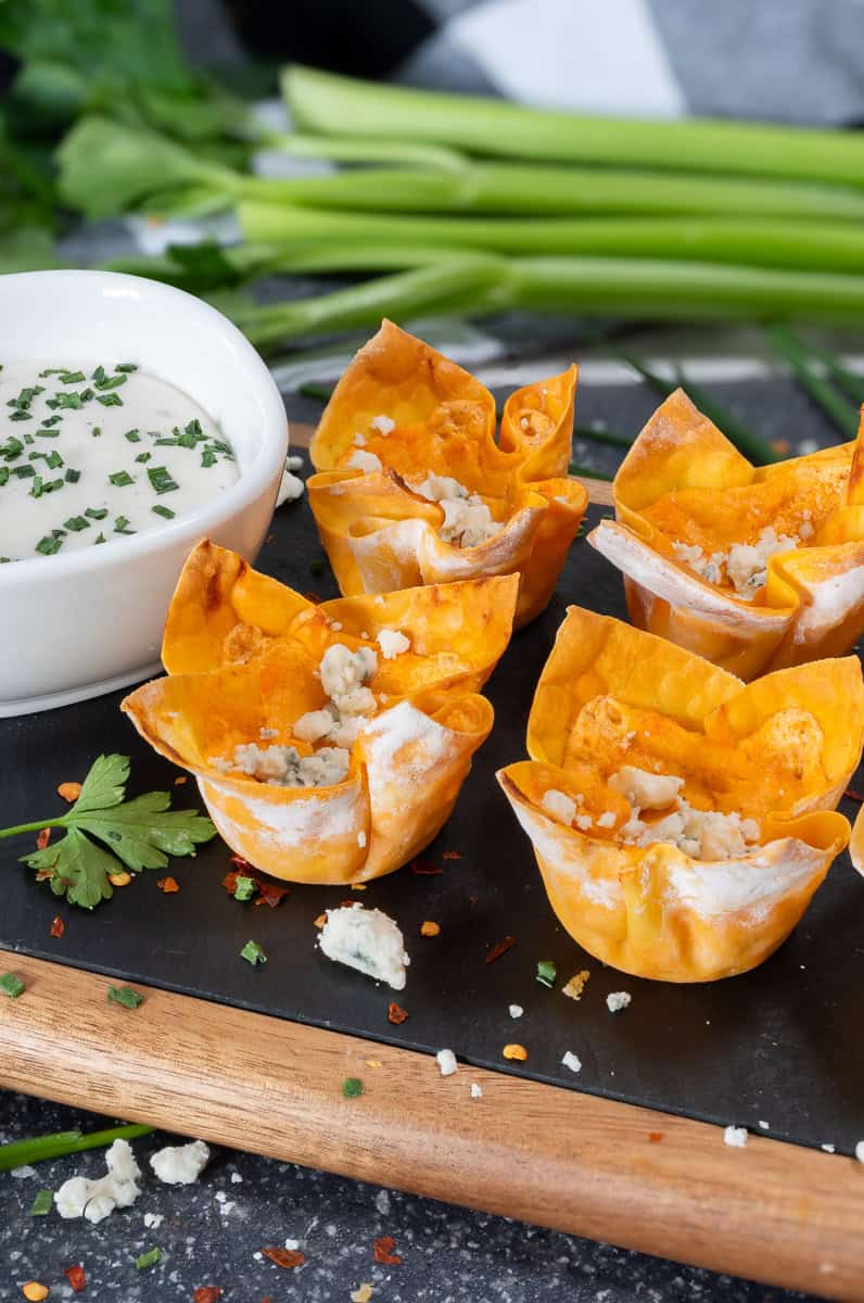 A platter with four crispy, golden-brown buffalo chicken wonton cups filled with a creamy mixture, garnished with blue cheese crumbles. A bowl of white dipping sauce with green herbs is next to the wonton cups, along with a garnish of parsley and chopped scallions in the background.