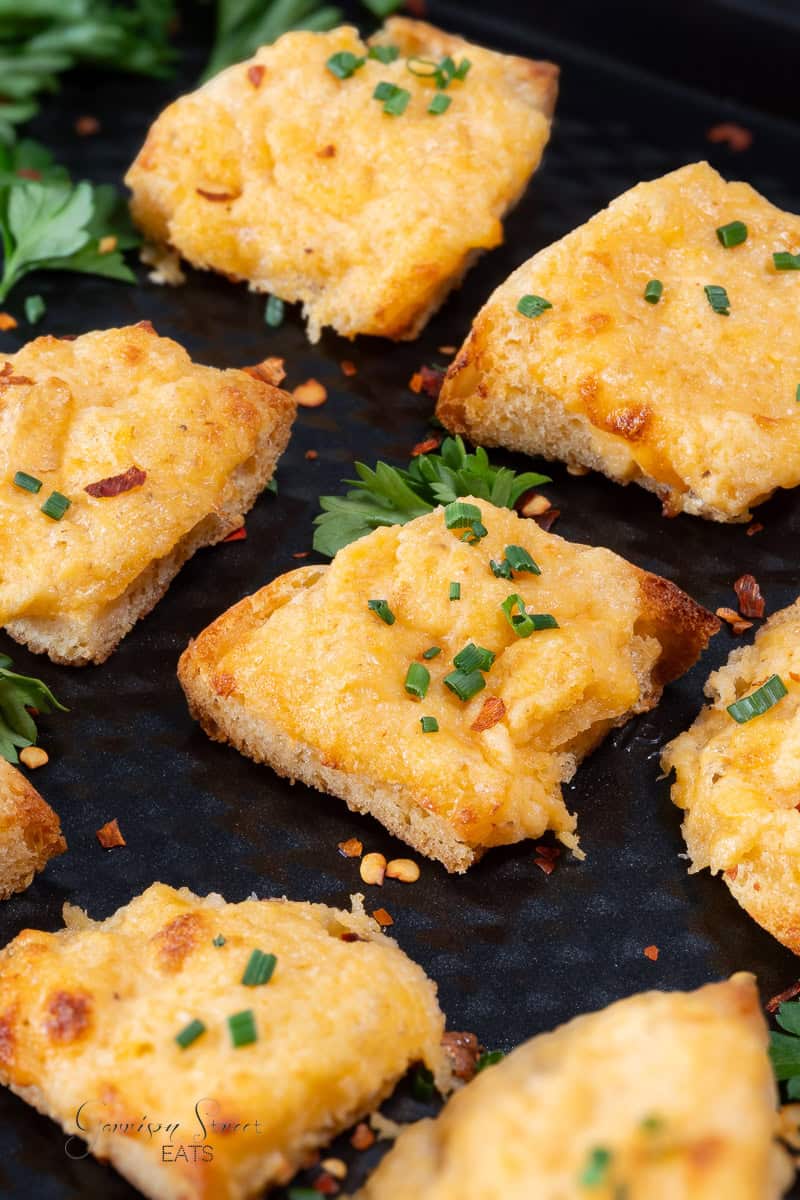 Close-up of several pieces of delectable crab appetizers on English muffins garnished with chopped chives, displayed on a dark, textured surface. In the background, sprigs of fresh parsley are slightly visible. The bread appears golden and crispy.