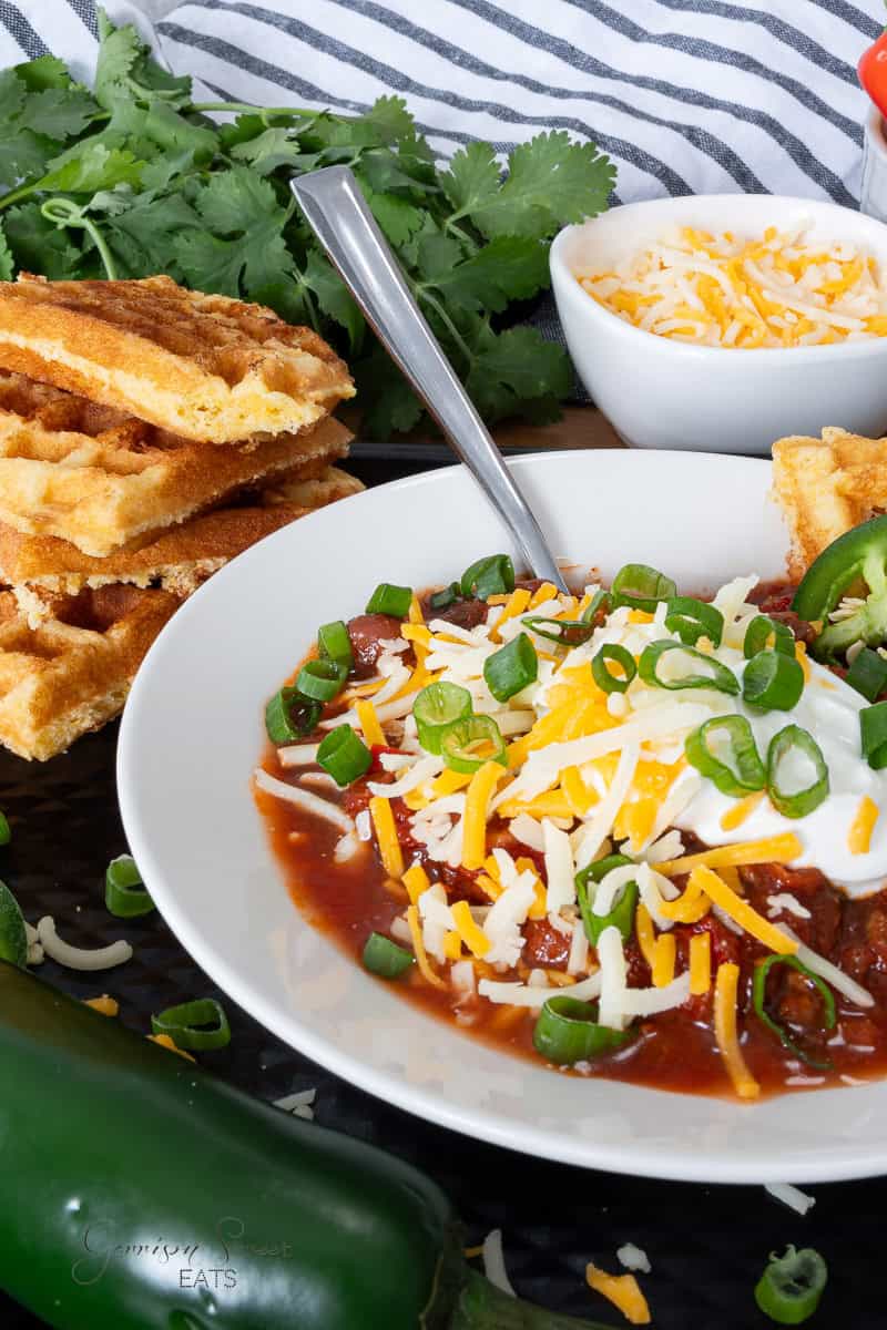 A bowl of chili topped with shredded cheese, sliced green onions, and sour cream sits on a table. The bowl is surrounded by a stack of cornbread waffles, cilantro leaves, a small bowl of shredded cheese, and a striped cloth. A jalapeño pepper is also visible in the foreground.