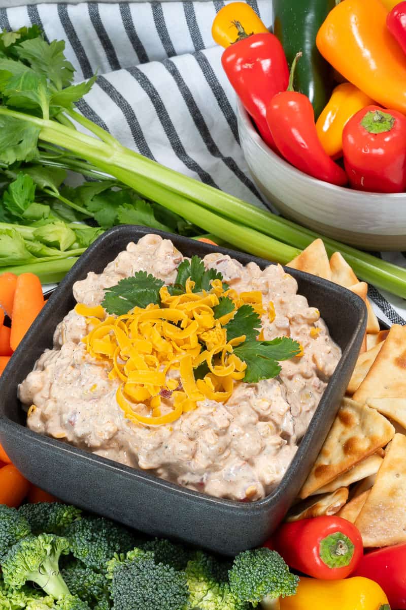 A black bowl containing a creamy Hidden Valley Ranch Fiesta Dip topped with shredded cheddar cheese and fresh cilantro. The bowl is surrounded by an assortment of fresh vegetables, including celery sticks, red and yellow mini bell peppers, carrot sticks, broccoli florets, and pita chips.