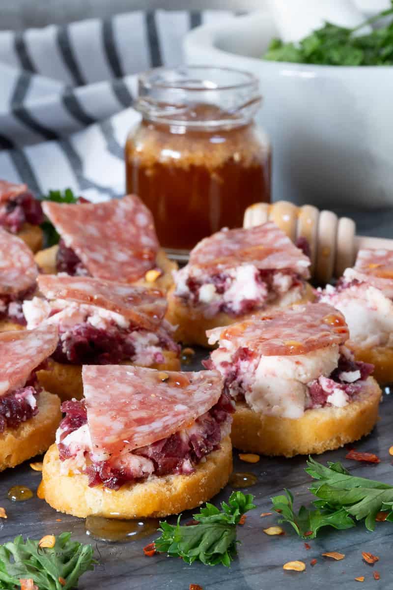 A close-up of several cranberry goat cheese crostini topped with thin slices of cured meat, drizzled with honey. Fresh herbs and a jar of honey with dipper are in the background, along with a striped napkin.