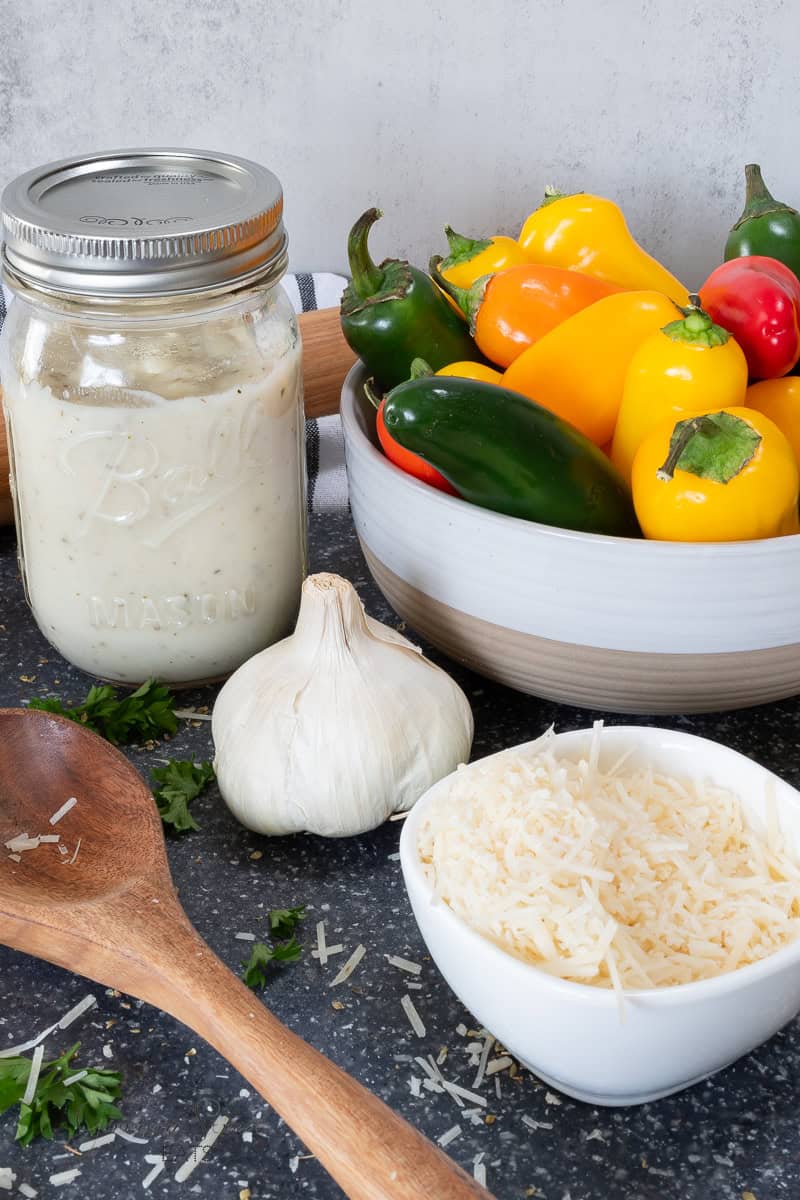 A kitchen counter with a jar of garlic pizza sauce, a bowl of assorted bell peppers, a garlic bulb, a bowl of grated cheese, and a wooden spoon. Fresh parsley leaves and cheese shavings are scattered on the dark countertop.