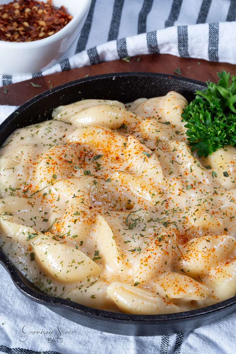 A skillet filled with creamy, cheesy shells and cheese, garnished with parsley and a sprinkling of paprika. A sprig of parsley is placed on the side, adding a touch of green. In the background, a small white bowl of crushed red pepper is visible, all on a striped cloth.