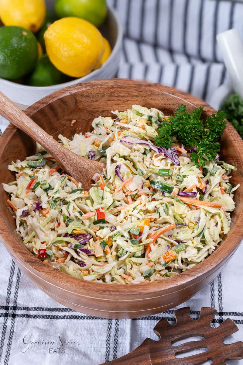 A wooden bowl filled with colorful coleslaw, including shredded cabbage, carrots, and red bell pepper pieces. A wooden spoon rests inside the bowl. Behind the bowl are lemons, limes, and a striped cloth. A sprig of parsley garnishes the coleslaw.