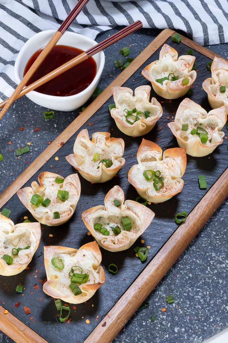 A wooden tray holds 10 crispy wonton wrapper appetizers garnished with chopped green onions. A small white bowl filled with dipping sauce and a pair of wooden chopsticks are placed at the top left of the tray. A black and white striped cloth is laid in the background.