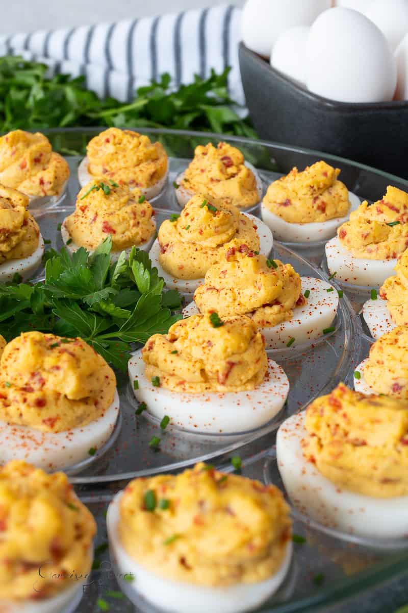 A platter of deviled eggs, topped with a vibrant yellow filling, garnished with red seasoning and chopped herbs. Fresh parsley decorates the center of the dish. A black bowl containing whole eggs and a striped cloth are in the background.