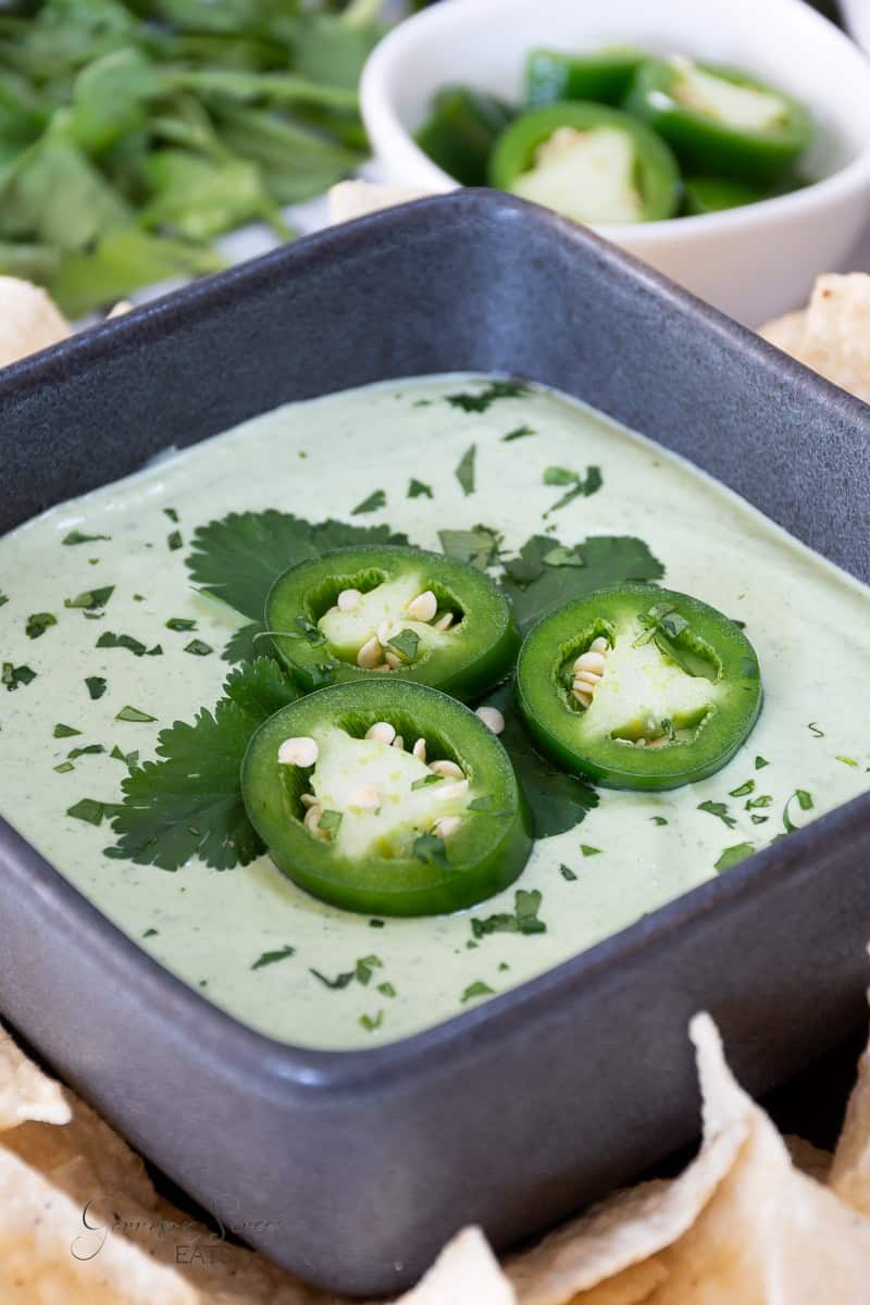 A square black bowl filled with creamy green chuys jalapeno dip, is topped with three slices of jalapeño peppers and garnished with cilantro. The bowl is surrounded by tortilla chips, and a small white dish with additional jalapeño slices is visible in the background.