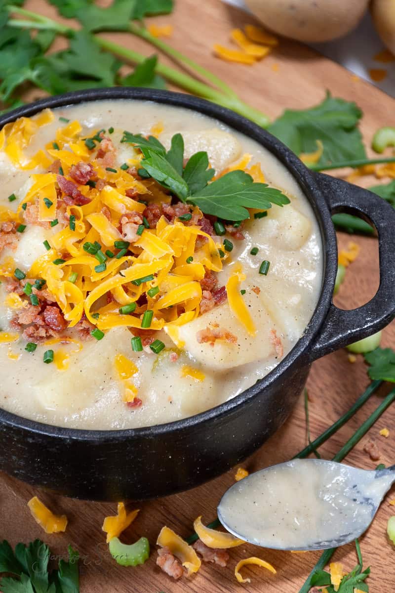 A black bowl filled with creamy, hearty, and chunky potato soup recipe garnished with shredded cheddar cheese, crispy bacon bits, chopped chives, and a sprig of parsley. A spoon with a bit of soup rests beside the bowl on a wooden surface scattered with greens and cheese.