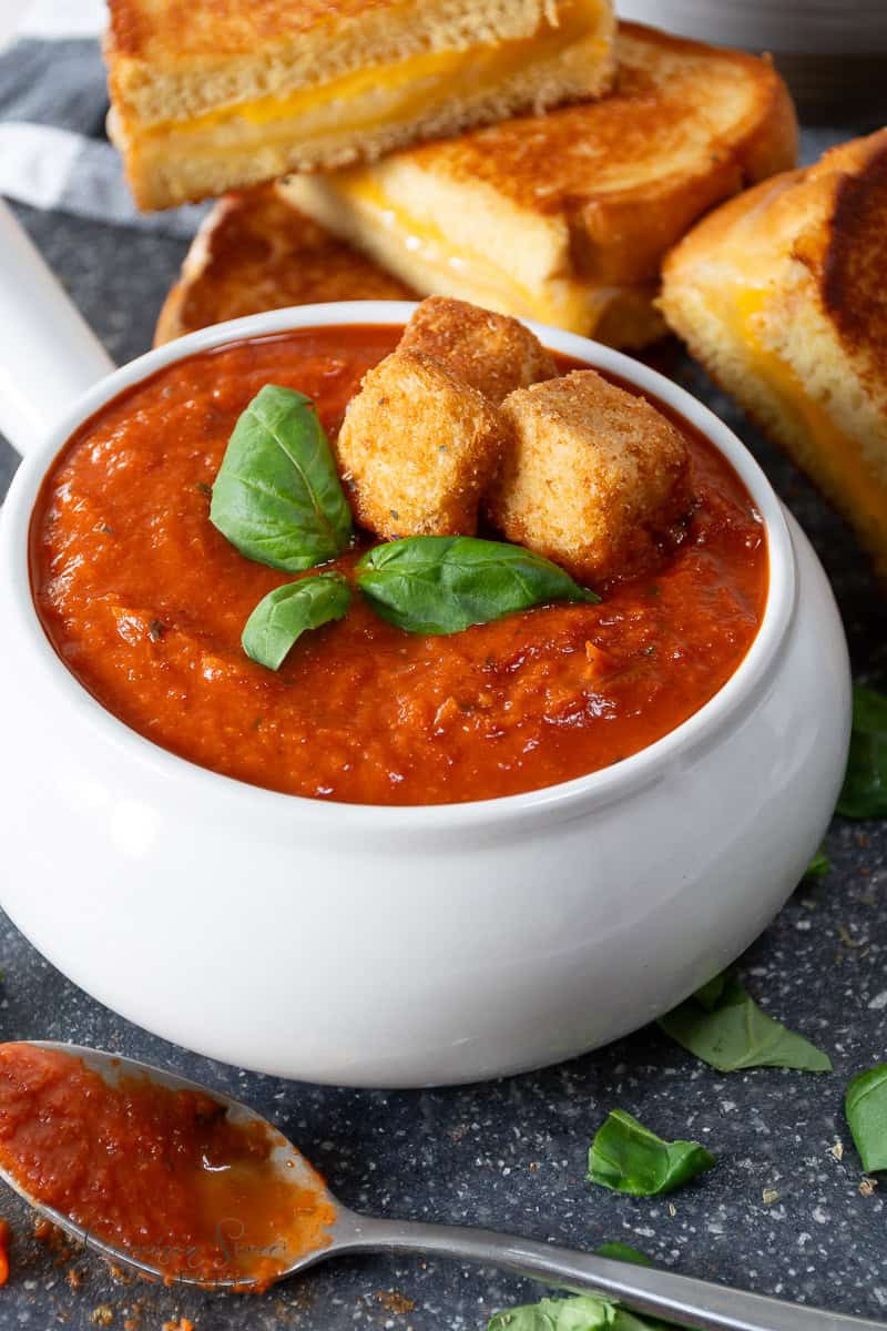 A bowl of creamy tomato basil soup garnished with fresh basil leaves and croutons, placed on a speckled countertop. In the background, there are grilled cheese sandwiches. A spoon with a bit of soup rests beside the bowl.