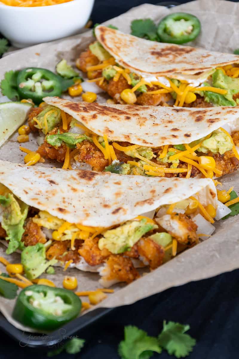 Grilled tortillas filled with shredded avocado, melted cheese, corn, and crispy chicken are laid out on parchment paper. Fresh cilantro, sliced jalapeños, and a lime wedge accompany the dish for a touch of zest.