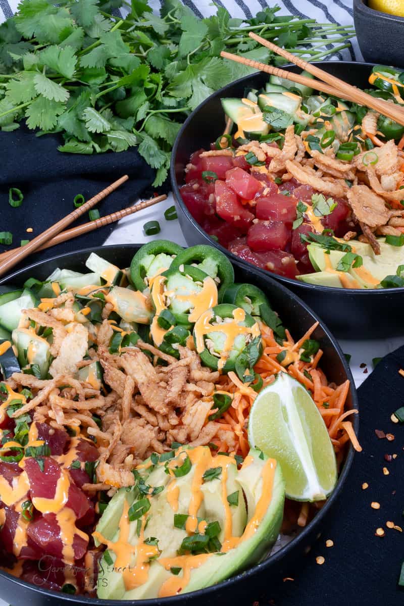 Two bowls of colorful poke topped with avocado slices, spicy tuna, shredded carrots, jalapeño slices, cucumber chunks, crispy onions, and a drizzle of sauce. Chopsticks rest on the bowl rims, and fresh cilantro leaves lie in the background.