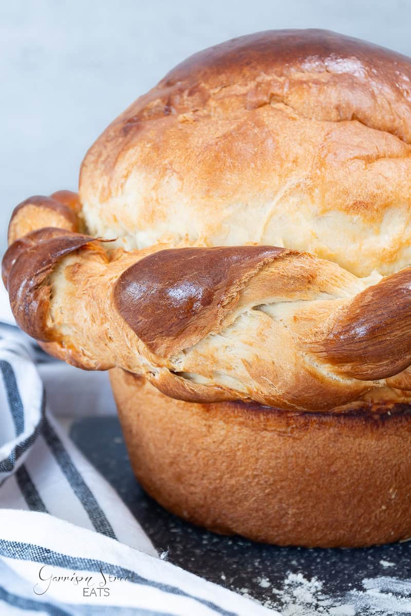 A close-up of the Easter bread—a golden-brown, braided loaf with a shiny surface. The tall, round bread sits on a dark surface next to a striped cloth napkin. Its texture is soft and fluffy, with a slightly glossy finish that invites you to take a bite.