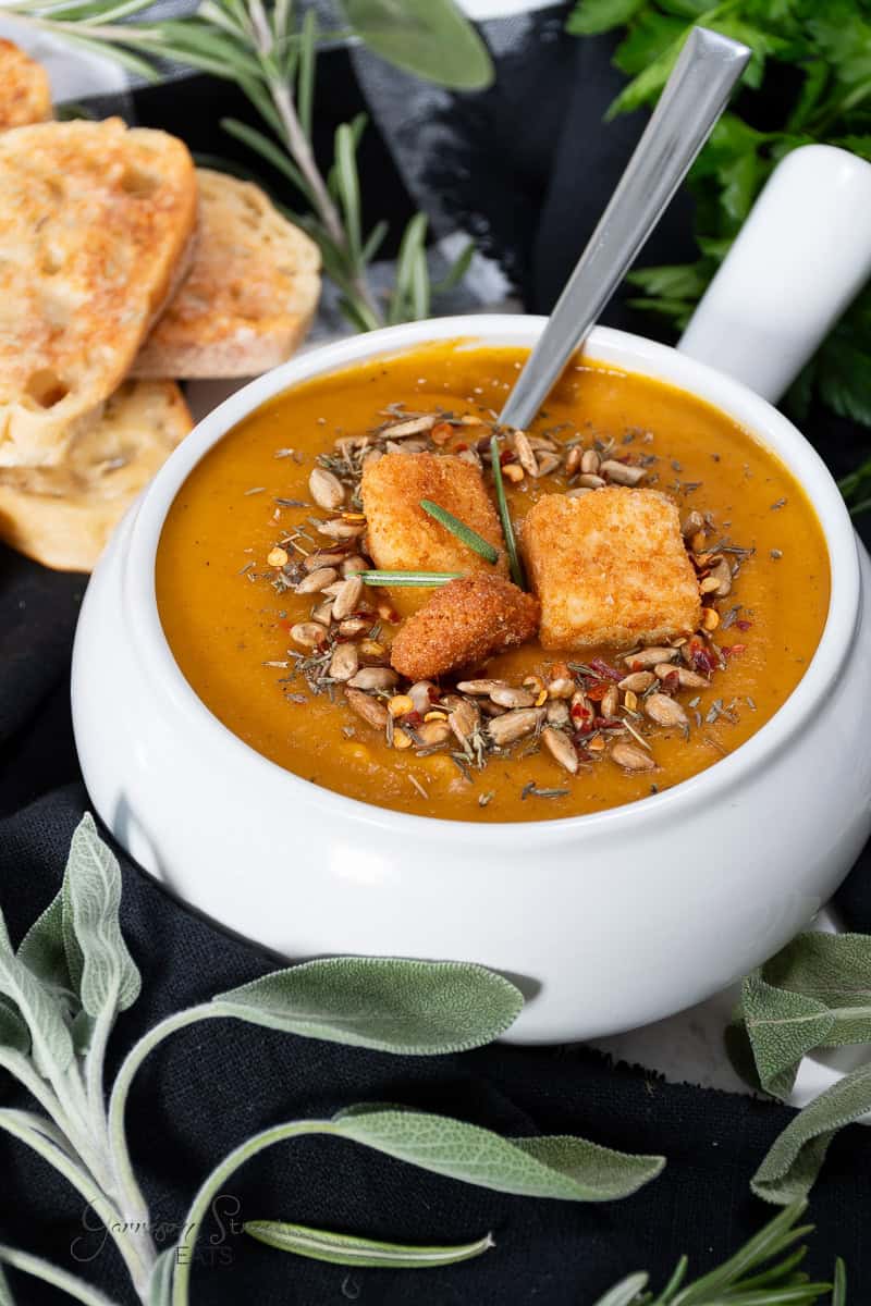A white bowl filled with creamy soup garnished with croutons, seeds, and fresh herbs. A spoon is placed in the bowl, and slices of toasted bread are in the background. Greenery, including fresh sage and rosemary, decorates the scene.