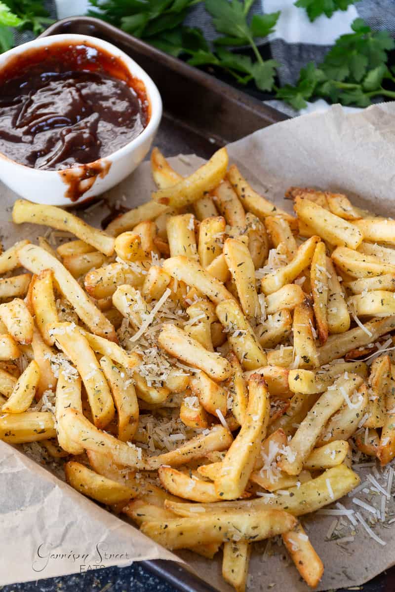 A tray of seasoned air fryer garlic fries topped with shredded cheese is shown on parchment paper. In the background, there is a small bowl of dark dipping sauce. Fresh parsley and a dark napkin are partially visible.