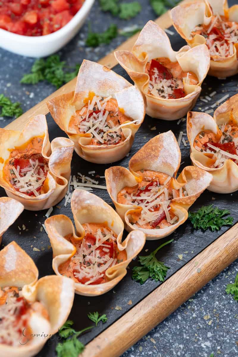 A tray of neatly arranged wonton pizza cups filled with melted cheese, small pepperoni slices, and topped with grated parmesan. The browned and crispy pepperoni appetizers are garnished with fresh parsley. A small bowl of diced tomatoes and herbs is visible in the background.