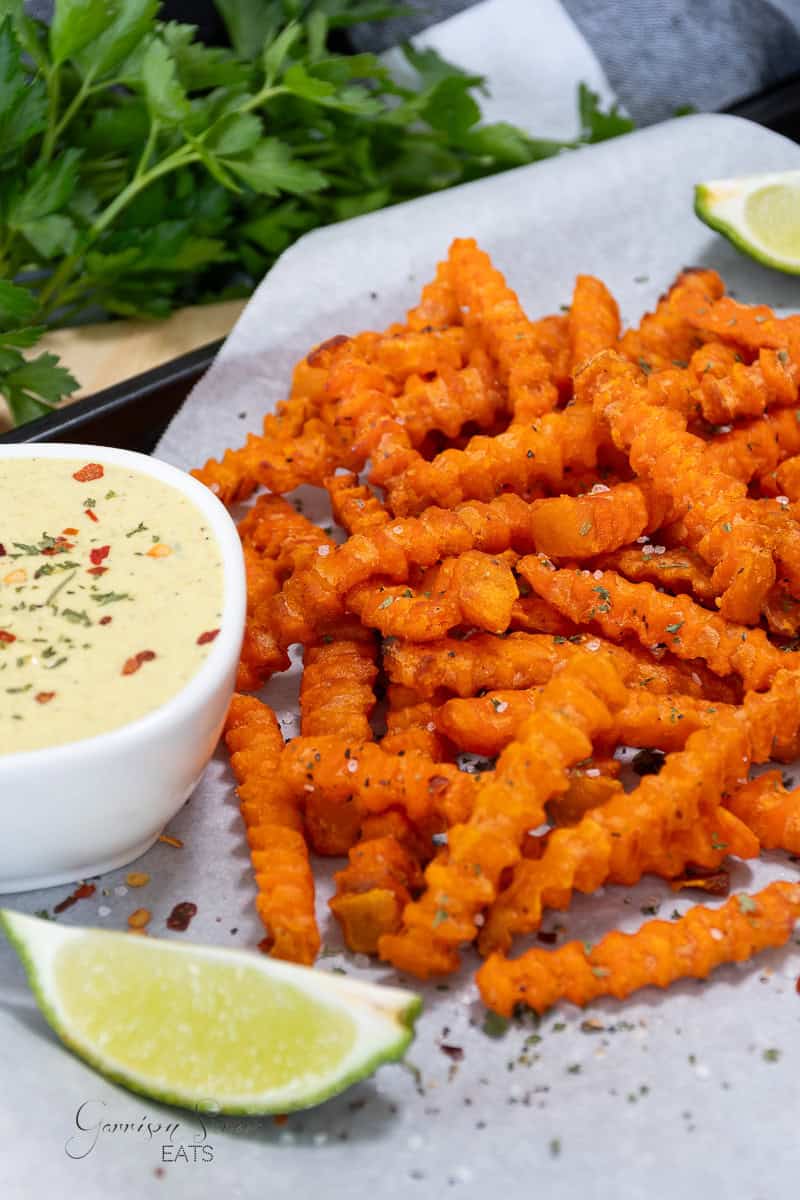 A plate of crispy, golden-orange crinkle-cut sweet potato fries cooked to perfection in a Ninja air fryer is served with a small bowl of creamy dipping sauce topped with herbs. Fresh parsley and lime wedges are placed nearby for garnish, and the fries are seasoned with pepper.