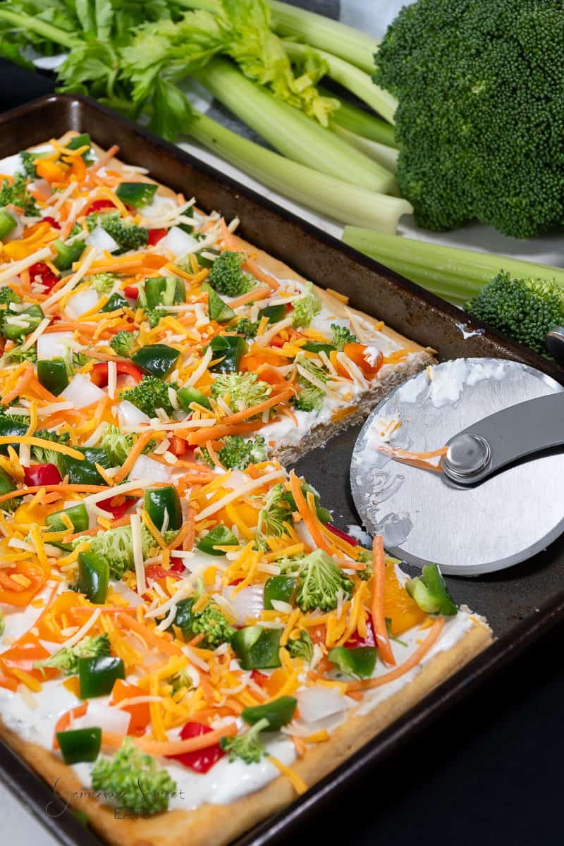 A rectangular Hidden Valley Ranch Veggie Pizza with Crescent Rolls is topped with colorful bell peppers, broccoli, and shredded cheese, displayed on a baking sheet. A slice has been cut out, and a pizza cutter with remnants of sauce is placed nearby. Fresh celery and broccoli are in the background.