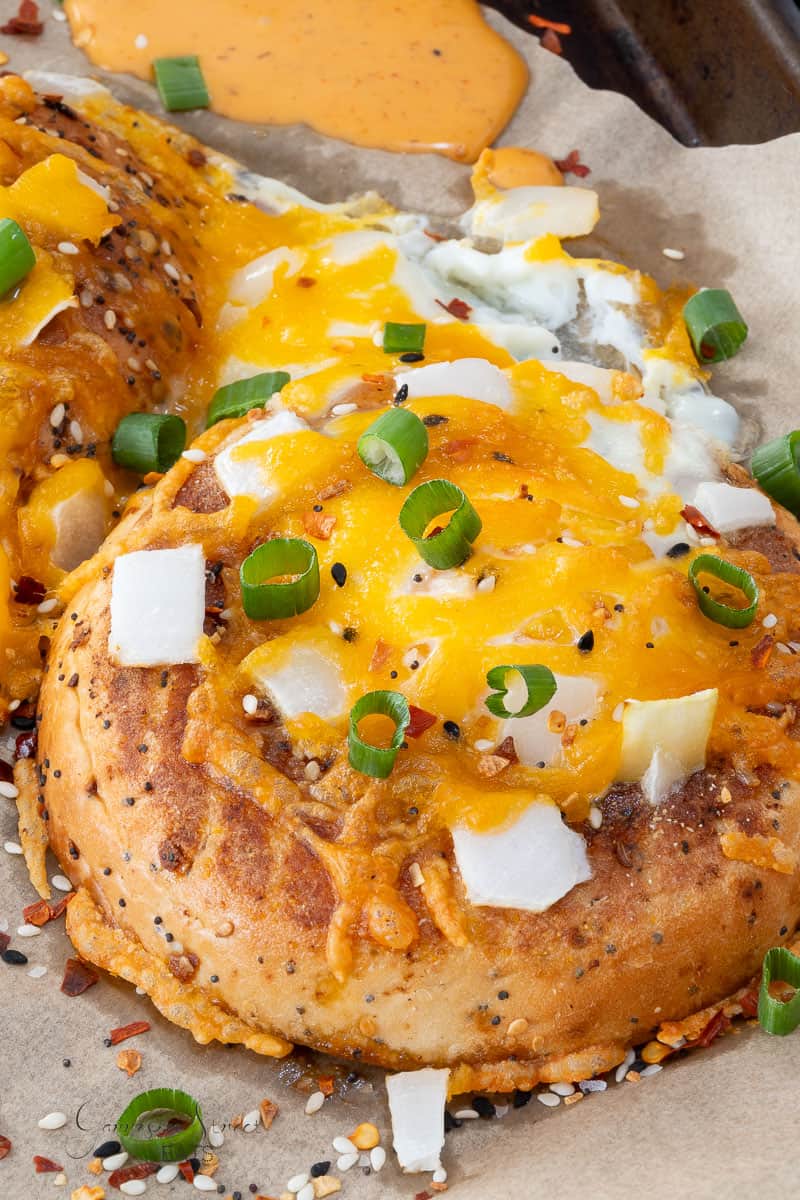 A close-up of a breakfast bagel split open with melted cheddar cheese, chopped green onions, white onion pieces, and seasoning on top. The bagel rests on parchment paper with a dollop of orange sauce in the background.