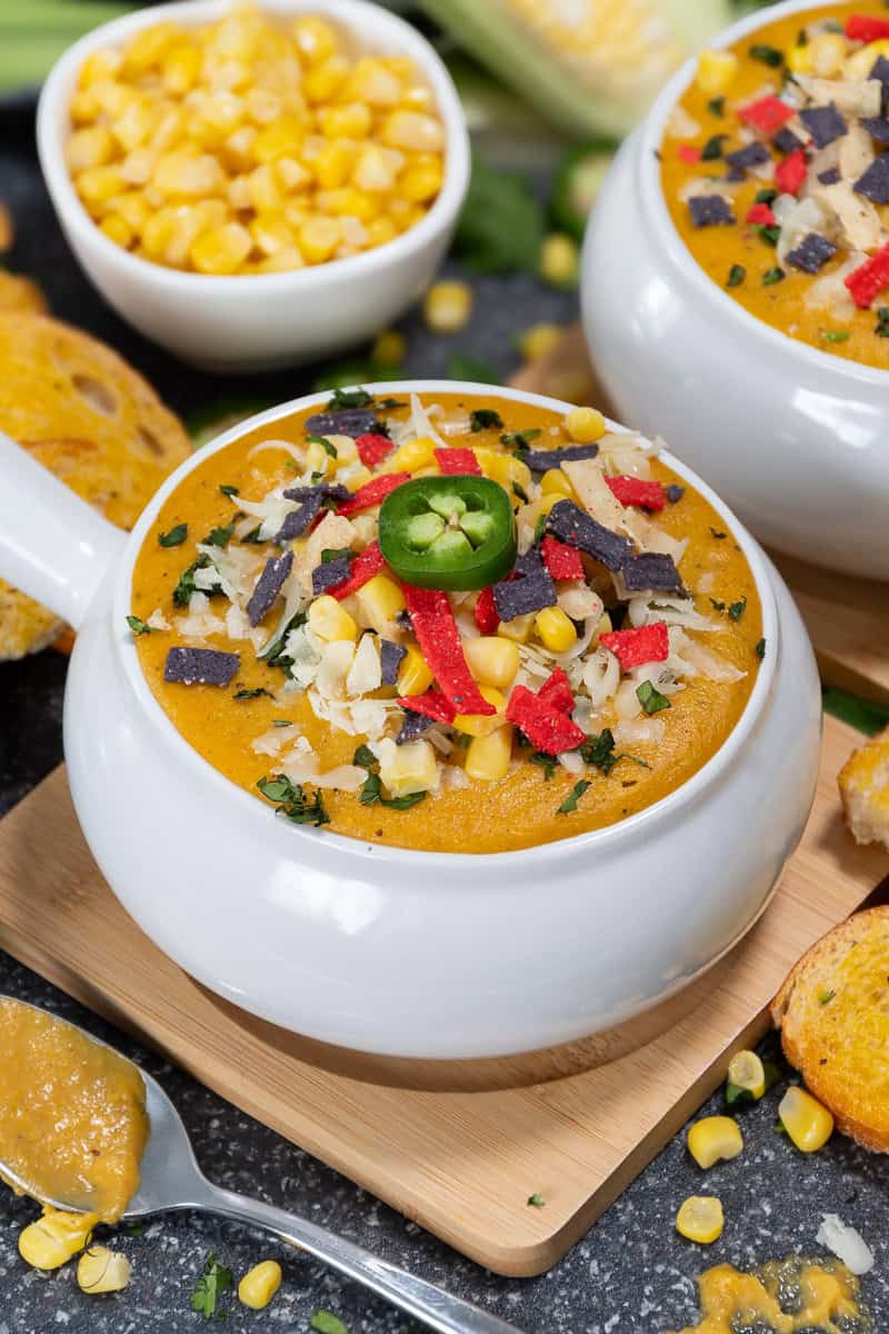 Two bowls of creamy corn soup garnished with shredded cheese, tortilla strips, and a slice of jalapeño. Corn kernels are scattered on the surface of the soup. In the background, a bowl of corn kernels and a slice of bread complete this sweet corn soup recipe. A spoon with some soup rests beside the bowl.