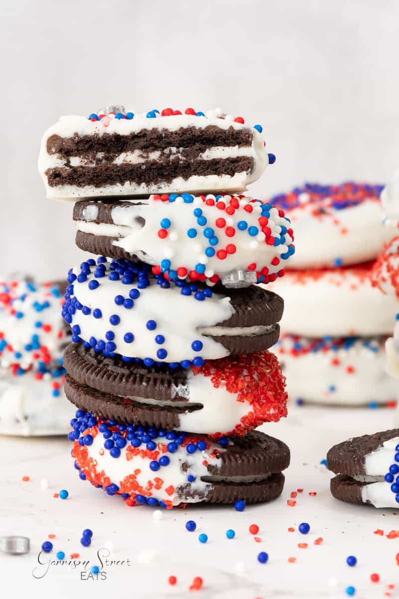 A stack of Oreo cookies dipped in white chocolate and adorned with red, white, and blue sprinkles makes for festive 4th of July cookies. One cookie on top is bitten, revealing the chocolate inside. Similar decorated cookies are blurred in the background.