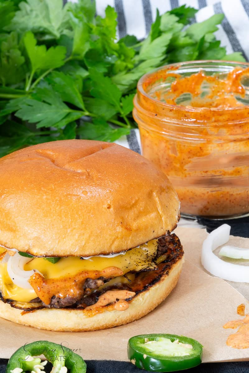 A juicy cheeseburger with melted cheese, onions, and sauce sits on parchment paper. Sliced jalapeños are nearby, and a jar of Copycat Kraft Chipotle Aioli is in the background. Fresh parsley leaves add a pop of green color to the scene.