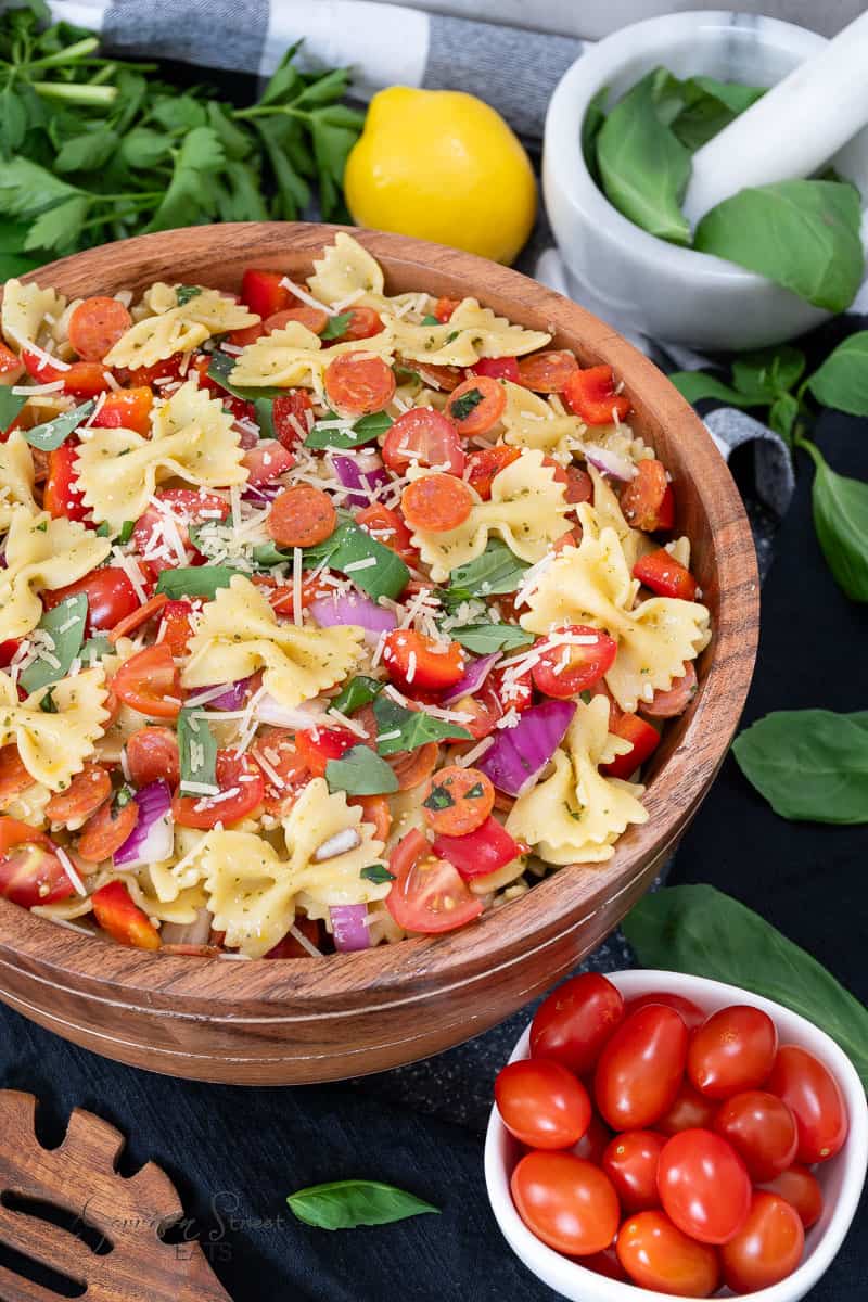 A vibrant pasta salad in a wooden bowl is filled with bowtie pasta, sliced cherry tomatoes, red onion, pepperoni slices, basil leaves, and topped with shredded Parmesan cheese. Fresh cherry tomatoes, basil, and a lemon are placed around the bowl.