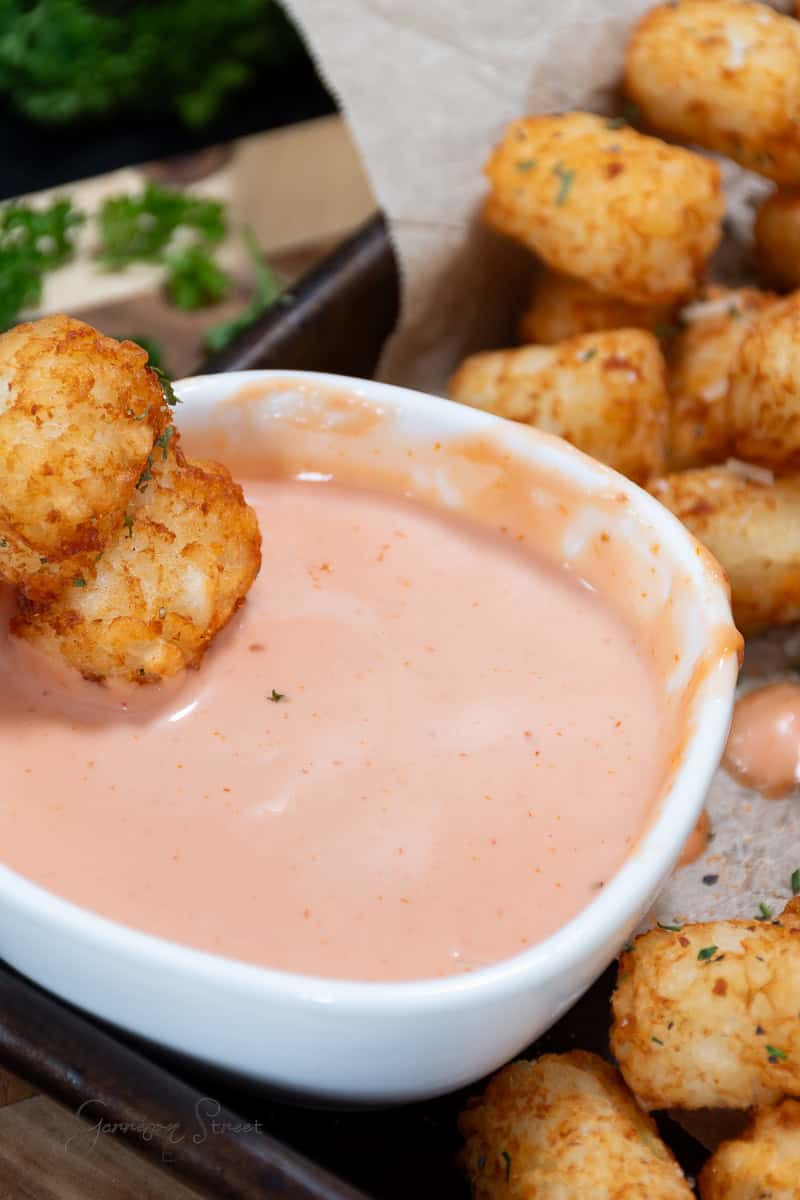 A close-up of a white bowl filled with pink mayo ketchup sauce, surrounded by golden, crispy tater tots. The bowl is placed on a tray lined with parchment paper, and a tater tot is being dipped into the sauce. Fresh green herbs are visible in the background.