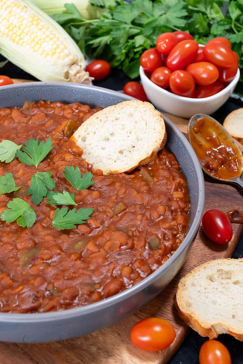 A hearty bowl of baked beans with bacon, garnished with fresh parsley and accompanied by slices of crusty bread. In the background, there are fresh cherry tomatoes in a bowl, ears of corn, and more parsley, all arranged on a wooden surface.