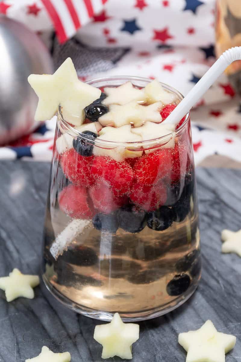 A festive white sangria recipe with vodka, served in a clear glass filled with blueberries, raspberries, and star-shaped apple slices. It's garnished with a star-shaped apple slice on the rim and a white straw. The backdrop features patriotic decor with red, white, and blue elements.