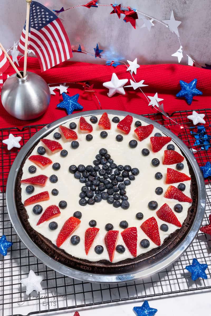 A festive dessert sits on a cooling rack. It is a round patriotic fruit pizza with a creamy white topping, decorated in a patriotic design with a star made of blueberries in the center, strawberry slices around the perimeter, and additional blueberries. Red, white, and blue decorations surround it.