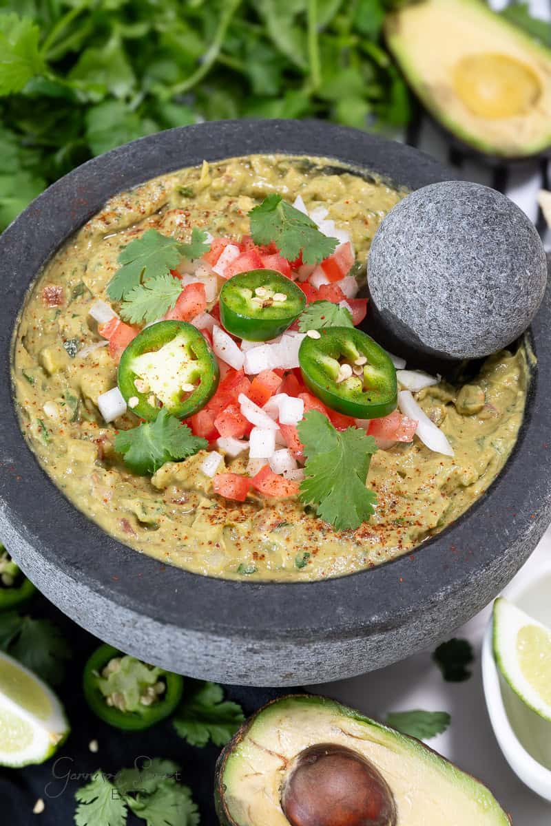 A stone bowl filled with guacamole garnished with diced tomatoes, onions, jalapeño slices, and cilantro leaves. The bowl is surrounded by fresh avocado, lime wedges, and more cilantro. A stone pestle rests on the edge of the bowl. Perfect for trying out a new jalapeno guacamole recipe!