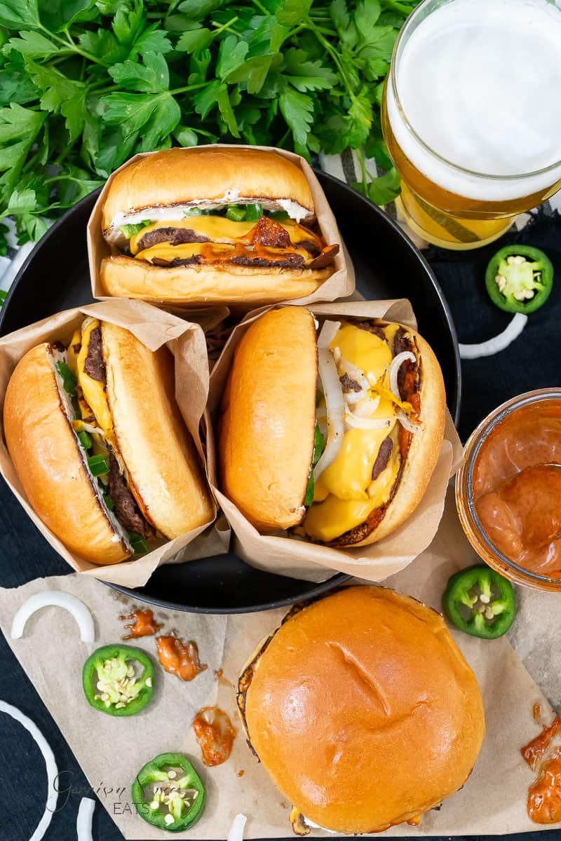 A top-view image of three cheeseburgers in paper wrappers on a black plate, garnished with slices of jalapeño and onions, with a jar of sauce beside them. A glass of beer and green herbs are in the background. Another smashburger from the Blackstone recipe is partially visible at the bottom.