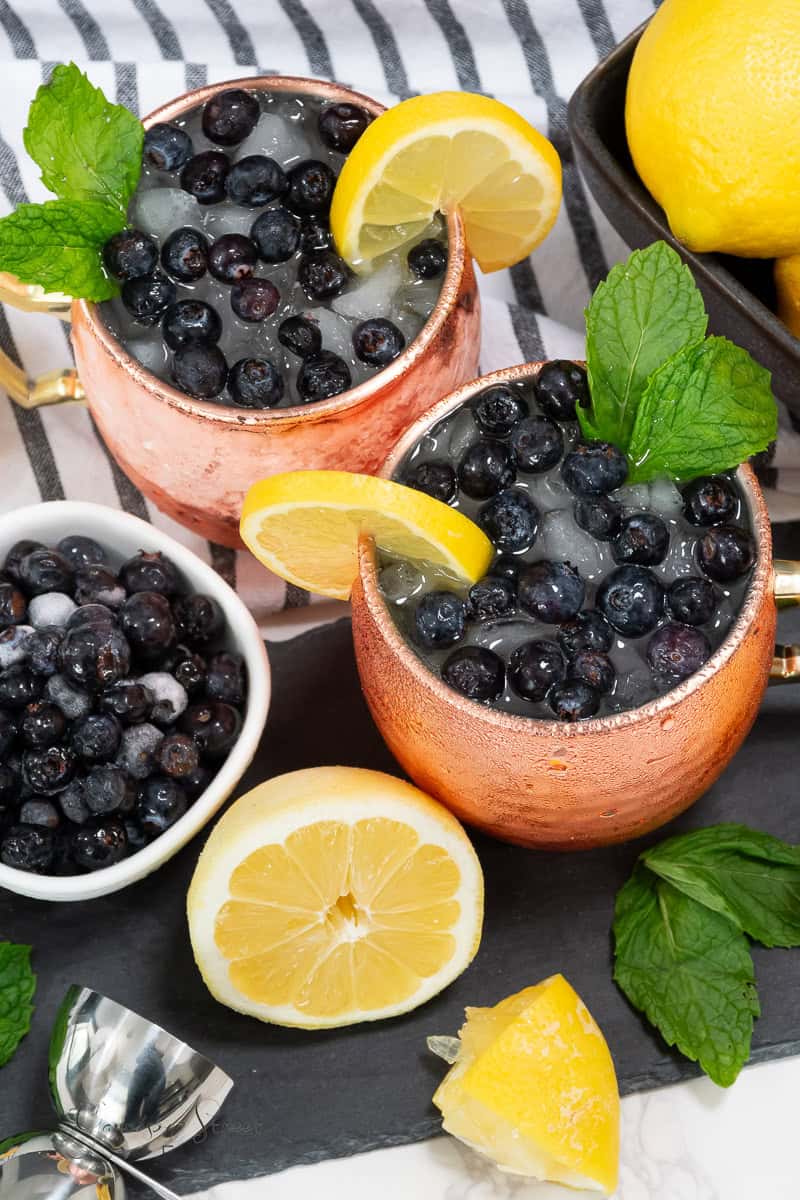 Two copper mugs filled with ice, blueberries, and garnished with lemon slices and mint leaves are placed on a striped cloth. A bowl of blueberries, a half and a whole lemon, a jigger, and additional mint leaves surround the mugs—perfect for your next blueberry vodka drinks recipe.