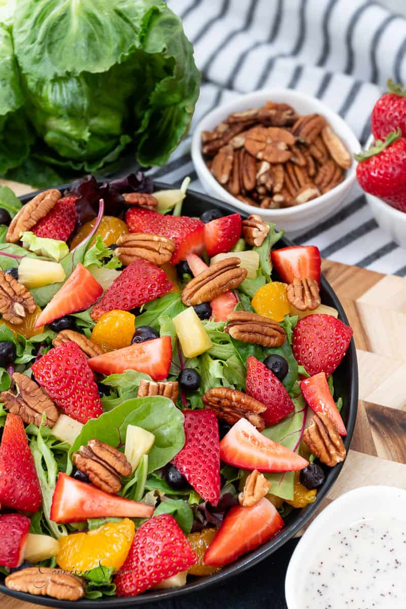 A fresh salad in a black bowl contains mixed greens, strawberries, blueberries, mandarin orange slices, pecans, and pineapple chunks. It is surrounded by a small bowl of pecans, a bowl of salad dressing, a head of lettuce, and whole strawberries.